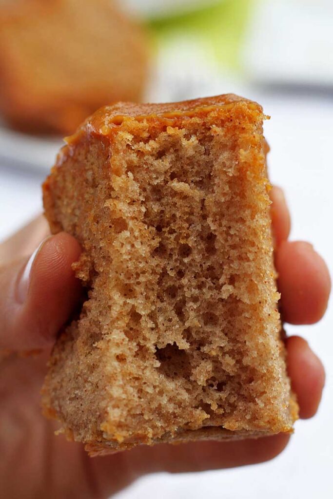Mão segurando fatia de bolo de canela com cobertura de doce de leite já mordido, mostrando a textura bem leve e aerada da fatia