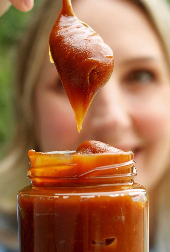 Mulher segurando pote e levantando uma colher de dentro cheia de doce de leite, a mulher está atrás do vidro desfocada
